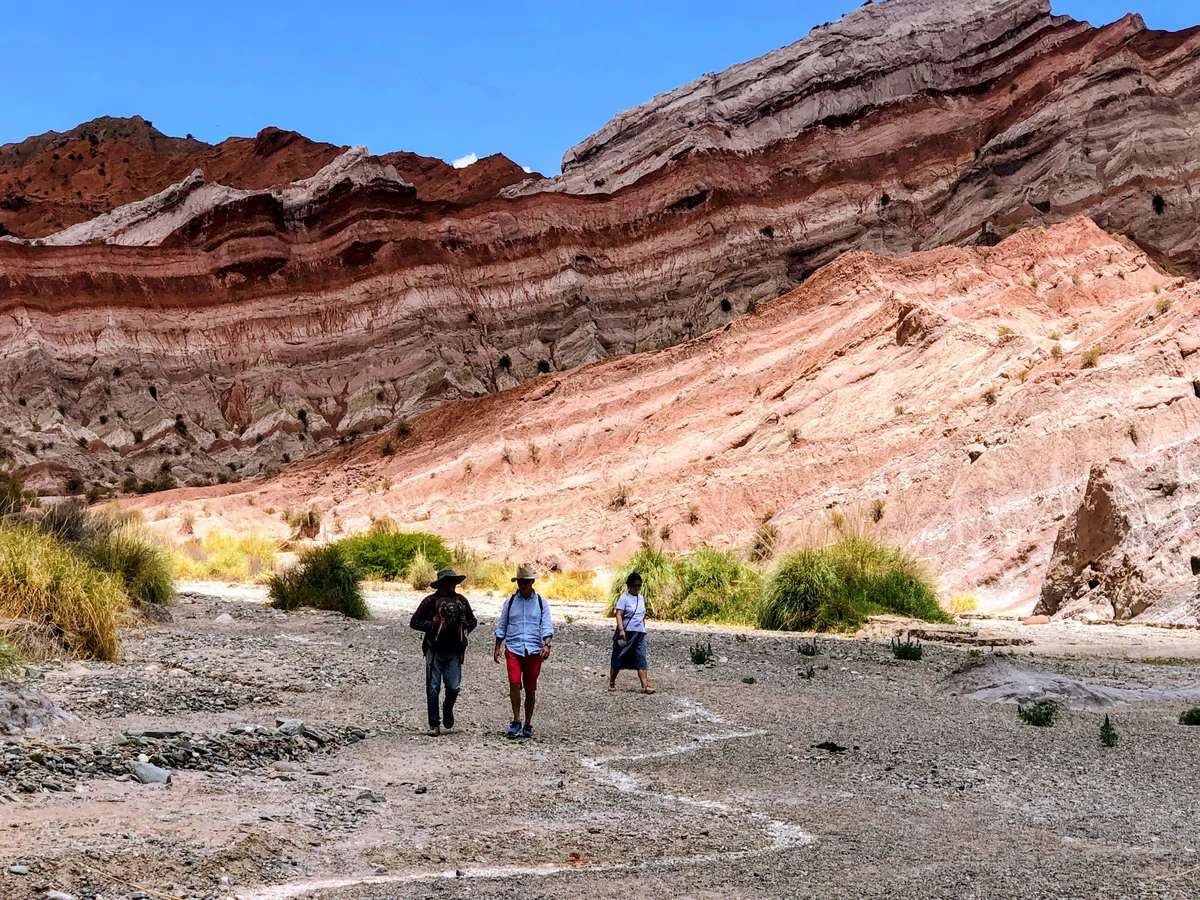 Three travellers walking under the striped red mountains of Jujuy